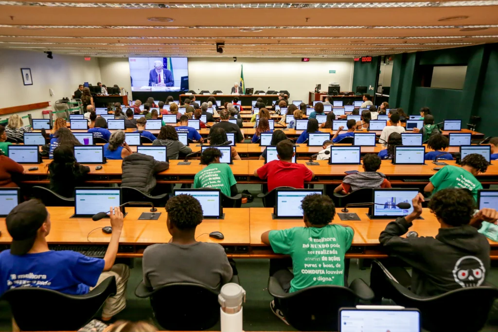 Rows of attendees of a forum in the Chamber of Deputies in Brazil sit at long tables in front of computer screens. 